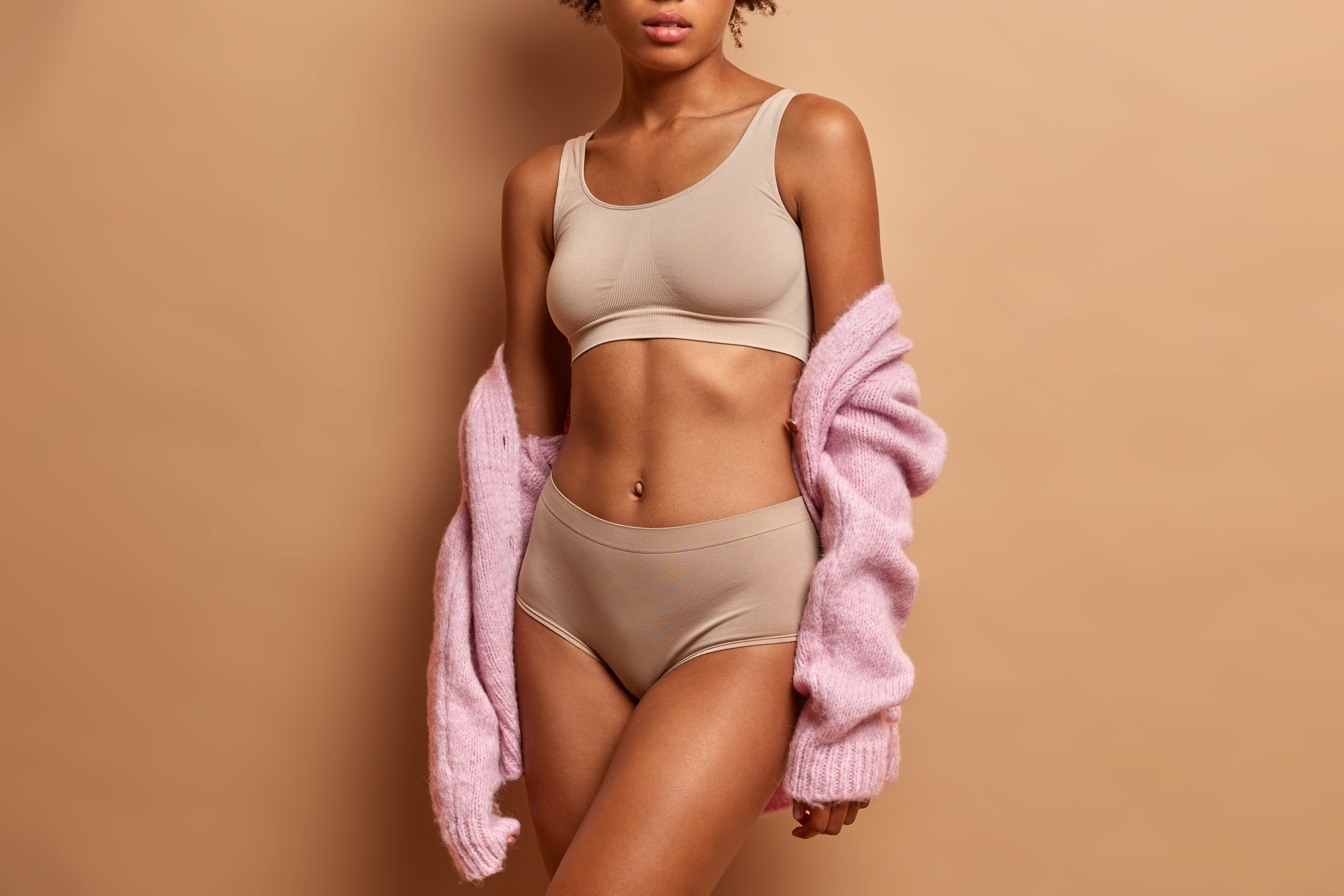 Unrecognizable slim young woman in lingerie has slim figure healthy skin perfect body shape poses against beige background. Studio shot.